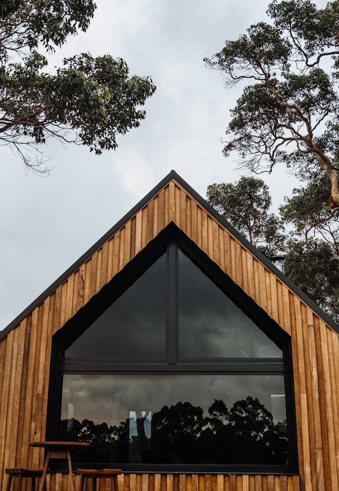 A stylish wooden cabin featuring a large triangular glass window, surrounded by trees.